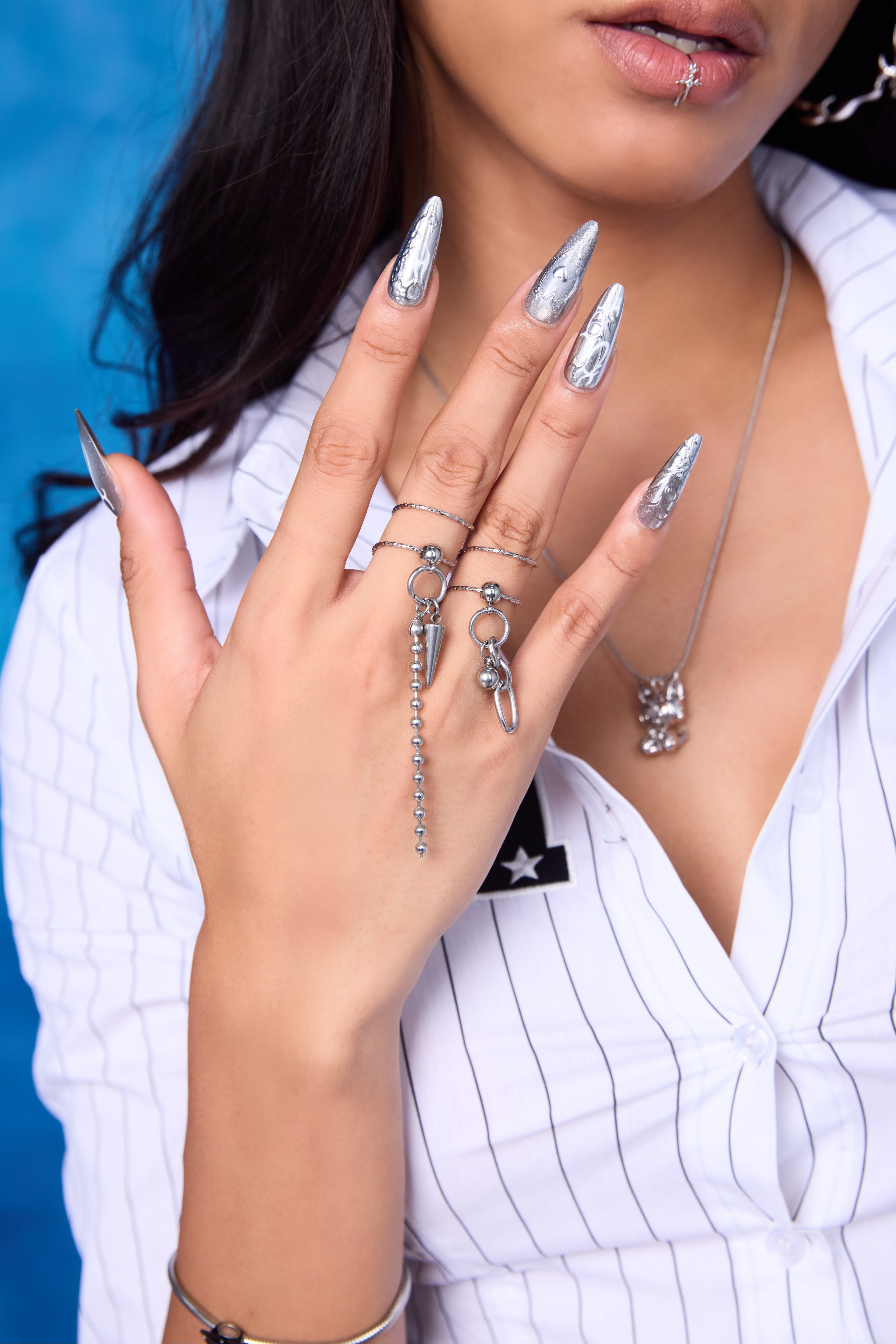 Riot Spike Ring Set on model — silver stacking rings with ball chain drop and spike pendant on middle finger, layered with slim band rings, chrome stiletto nails, FEFE Dog Necklace visible at chest, blue backdrop