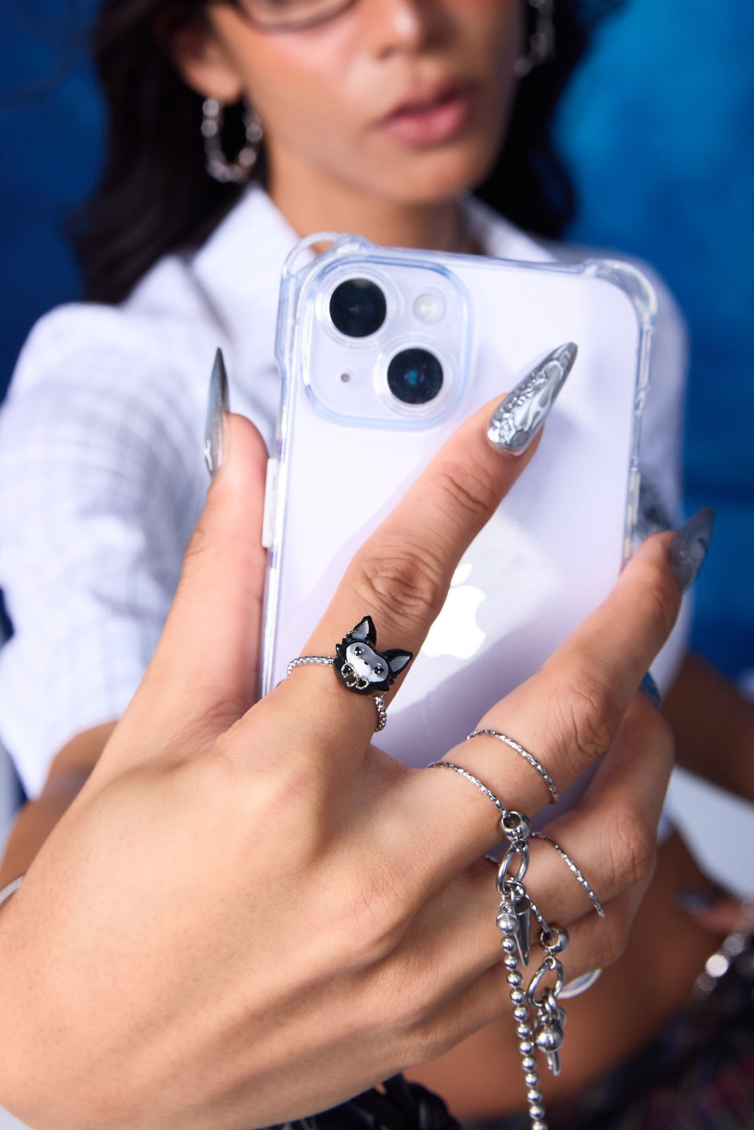 FEFE Dog Ring on model holding phone — adjustable silver ring with FEFE dog face charm on index finger, stacked with slim silver rings and ball chain drop, chrome nails, blue backdrop